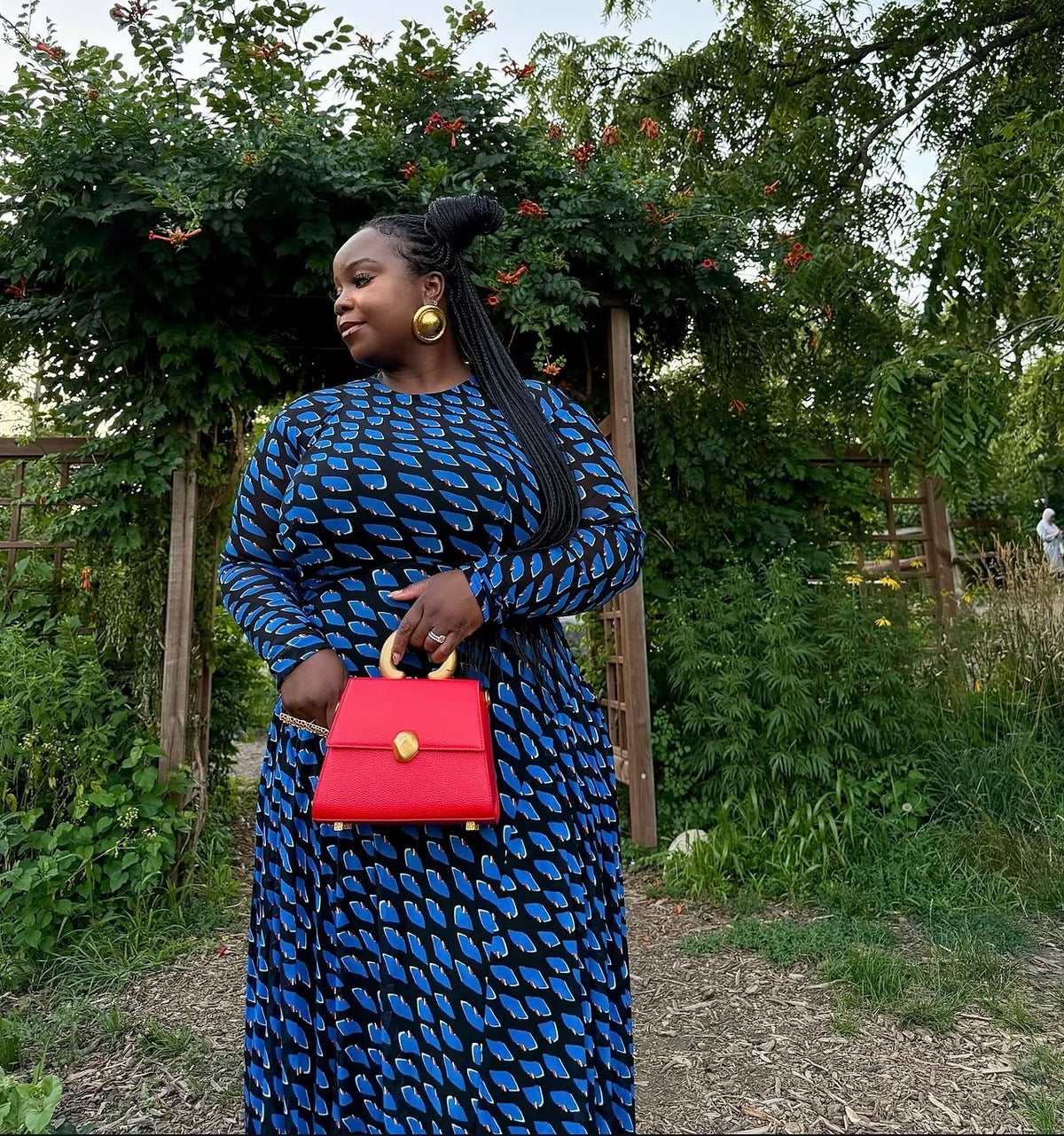 Woman wearing round gold Esinam earrings with a blue patterned dress outdoors.